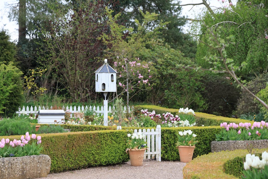 A well-maintained formal garden with lush green grass and neatly edged flower beds featuring a variety of colorful plants and flowers, including pinks, purples, and whites. In the foreground, a circul